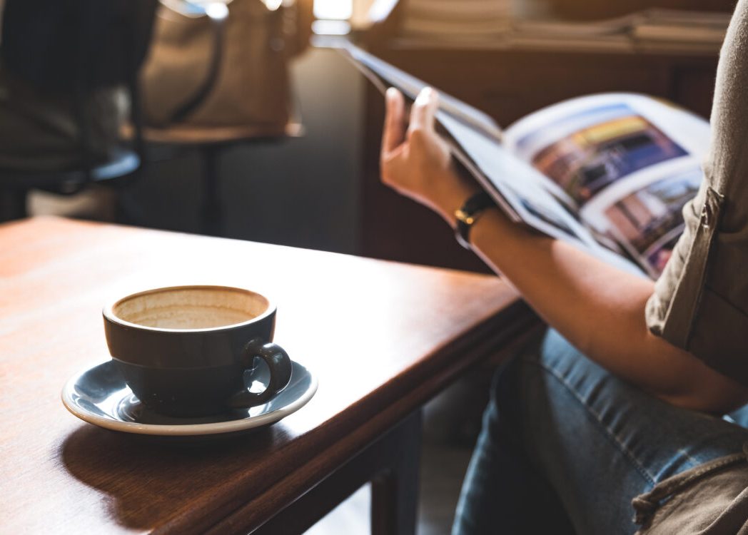 Closeup image of a woman reading a book with coffee cup on wooden table in modern cafe