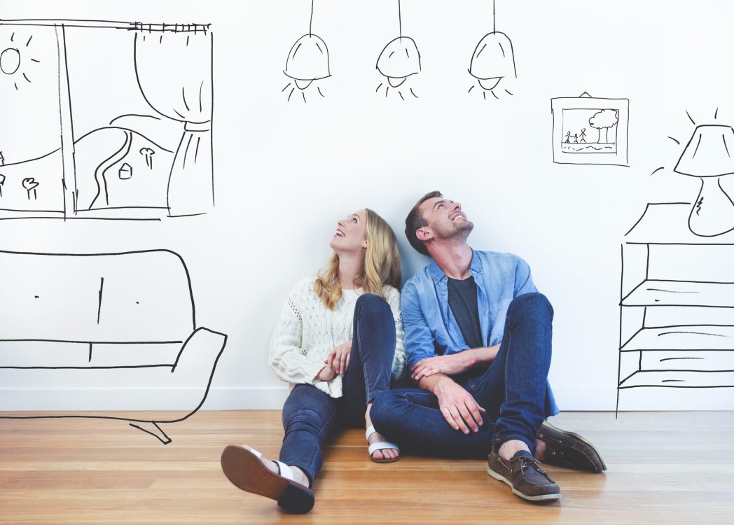 Couple dreaming of their new house. They are sitting on a wooden floor imagining their new home with furniture in it. The house is currently empty. They are booth looking up and smiling happily.